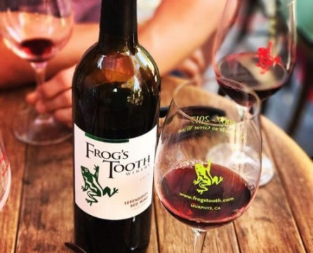 bottle of Frog's Tooth wine and glass of wine with the winery logo on a wine barrel table top